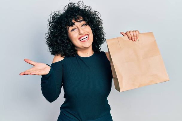 Young middle east woman holding take away paper bag celebrating achievement with happy smile and winner expression with raised hand  - 写真・画像