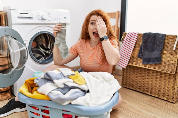 Mujer pelirroja joven poniendo ropa sucia en la lavadora que cubre un ojo con la mano, sonrisa confiada en la cara y emoción sorpresa.  - Foto, imagen