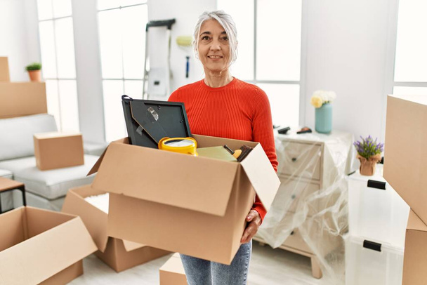 Mujer de pelo gris de mediana edad sonriendo confiado sosteniendo paquete en el nuevo hogar - Foto, imagen