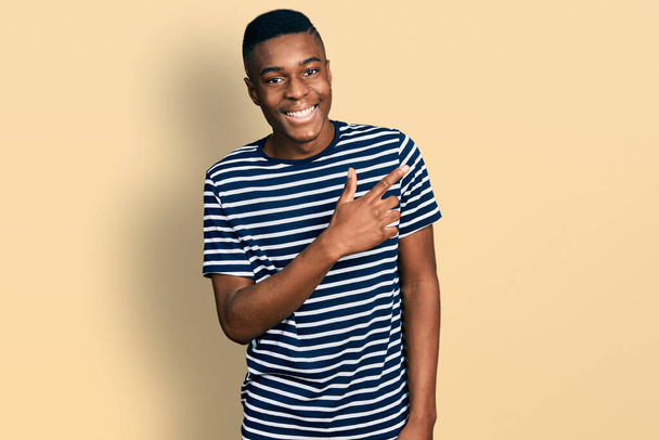 Mladý africký Američan muž na sobě ležérní pruhované tričko s veselým úsměvem ukazuje rukou a prstem na stranu  - Fotografie, Obrázek