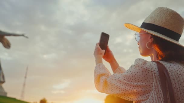 Une jolie femme près de Mère Patrie prenant une photo sur un smartphone au coucher du soleil - Séquence, vidéo