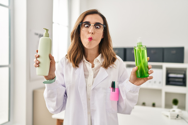 Young doctor woman holding aloe vera and cream making fish face with mouth and squinting eyes, crazy and comical.  - Foto, afbeelding