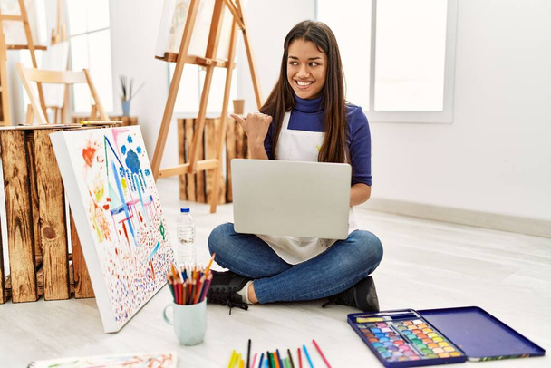Молодая брюнетка сидит на полу в художественной студии с ноутбуком указывая пальцем вверх в сторону улыбается счастливо с открытым ртом  - Фото, изображение
