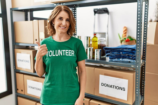 Keski-ikäinen nainen yllään vapaaehtoinen t paita lahjoituksia seistä hymyillen onnellinen ja positiivinen, peukalo ylös tekee erinomaista ja hyväksymismerkki  - Valokuva, kuva