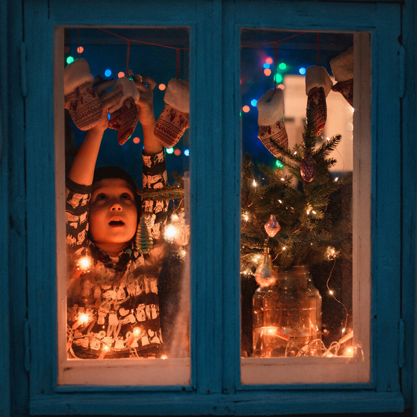 Sonriente niña dulce de pie junto a la ventana en tiempo de Navidad. Con luces de colores del árbol de Navidad en el fondo, enfoque selectivo. - Foto, imagen