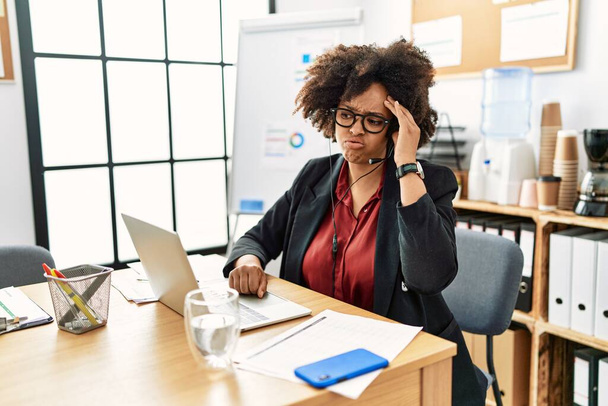 Femme afro-américaine avec des cheveux afro travaillant au bureau portant casque opérateur inquiet et stressé par un problème avec la main sur le front, nerveux et anxieux pour la crise  - Photo, image