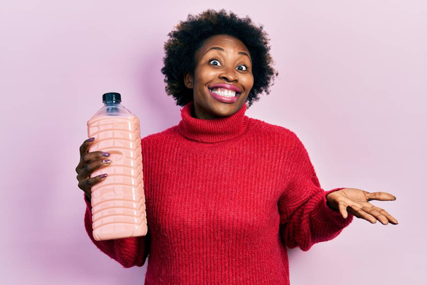 Giovane donna afroamericana in possesso di bottiglia detergente celebrando il raggiungimento con sorriso felice e l'espressione del vincitore con mano alzata  - Foto, immagini
