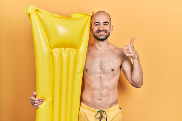 Молодий лисий чоловік в купальнику і тримає літній матрац плаває усміхаючись щасливим і позитивним, великий палець робить відмінний знак схвалення
  - Фото, зображення