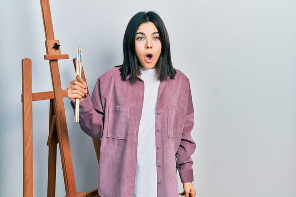 Mladá brunetka žena malíř sedí v ateliéru umění drží umělecké slovo vyděšený a ohromen s otevřenými ústy pro překvapení, nevěřící tvář  - Fotografie, Obrázek