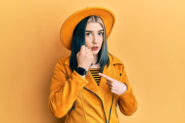 Jeune fille moderne portant chapeau jaune et veste en cuir pressé pointant vers le temps de montre, impatience, regardant la caméra avec une expression détendue  - Photo, image