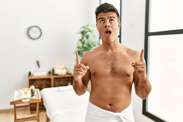 Young hispanic man standing shirtless at spa center amazed and surprised looking up and pointing with fingers and raised arms.  - Foto, immagini