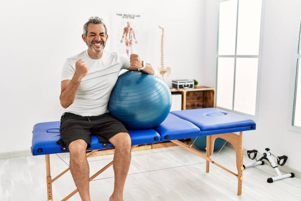 Middle age hispanic man at pain recovery clinic holding pilates ball very happy and excited doing winner gesture with arms raised, smiling and screaming for success. celebration concept.  - Photo, Image