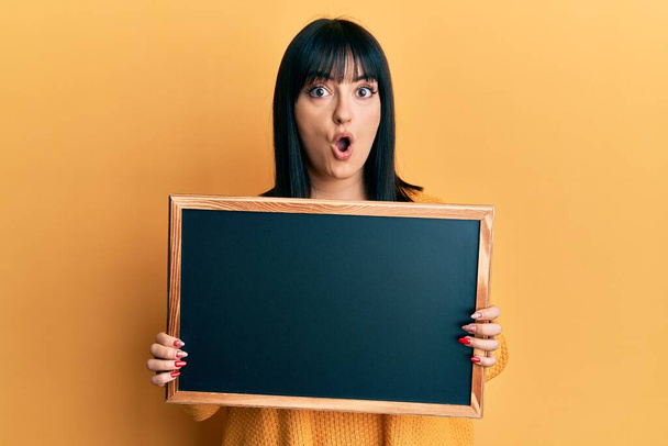 Young hispanic woman holding blackboard afraid and shocked with surprise and amazed expression, fear and excited face.  - Photo, Image