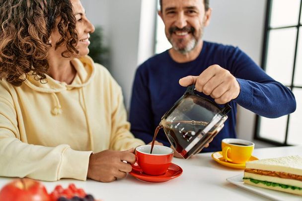 Latynoska para w średnim wieku uśmiechnięta szczęśliwa siedząca na stole jedząca śniadanie w domu. - Zdjęcie, obraz