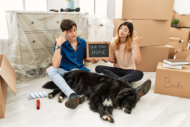 Young caucasian couple with dog holding our first home blackboard at new house shooting and killing oneself pointing hand and fingers to head like gun, suicide gesture.  - Photo, image