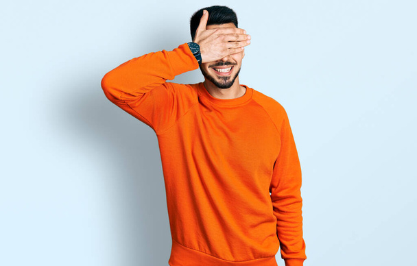 Junger hispanischer Mann mit Bart, lässig in orangefarbenem Pullover, lächelnd und lachend mit der Hand auf dem Gesicht, die Augen zur Überraschung verdeckt. Blindes Konzept.  - Foto, Bild