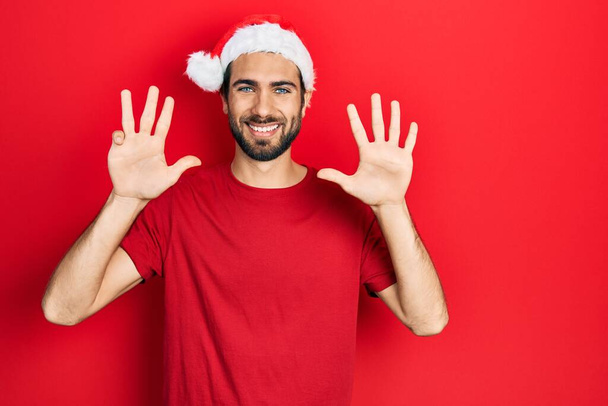 De jonge Spaanse man met de kerstmuts op en wijzend met vingers nummer negen terwijl hij zelfverzekerd en gelukkig glimlacht.  - Foto, afbeelding