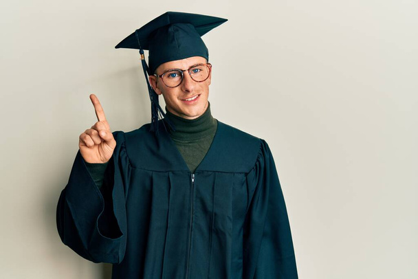 Young caucasian man wearing graduation cap and ceremony robe showing and pointing up with finger number one while smiling confident and happy.  - Photo, Image