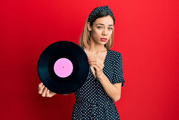 Young caucasian blonde woman holding vinyl disc clueless and confused expression. doubt concept.  - Photo, Image