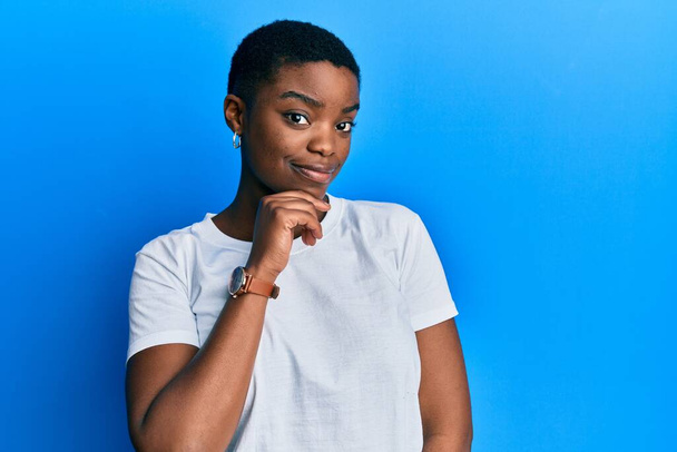 カジュアルホワイトのTシャツを着た若いアフリカ系アメリカ人女性が、交差する腕と顎の手でカメラに自信を持って笑っています。ポジティブな.  - 写真・画像