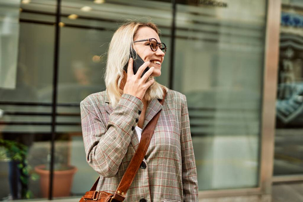Junge blonde Geschäftsfrau lächelt glücklich und plaudert auf dem Smartphone in der Stadt. - Foto, Bild