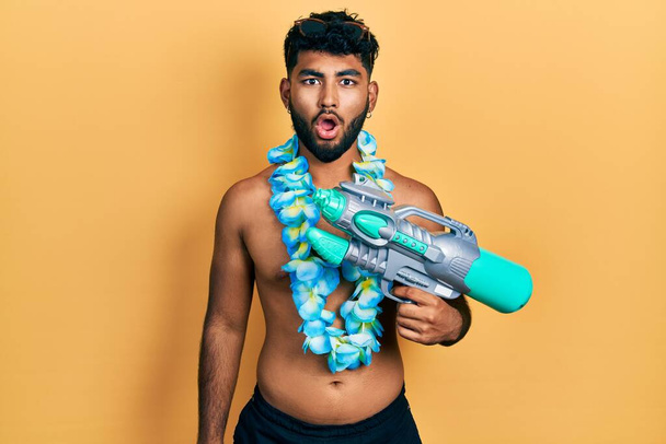 Homem árabe com barba vestindo maiô e hawaiian lei segurando watergun assustado e espantado com a boca aberta para surpresa, rosto descrença  - Foto, Imagem