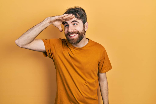 Uomo caucasico con la barba indossa casual t shirt gialla molto felice e sorridente guardando lontano con mano sopra la testa. concetto di ricerca.  - Foto, immagini