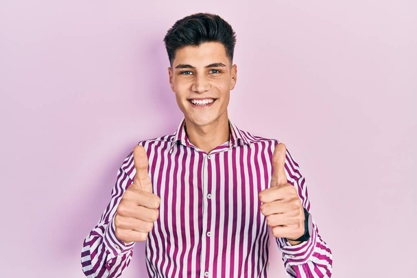 Joven hombre hispano usando ropa casual aprobando hacer gesto positivo con la mano, pulgares arriba sonriendo y feliz por el éxito. gesto ganador.  - Foto, Imagen