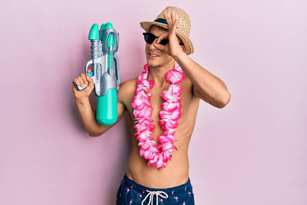 Young caucasian man wearing swimsuit and hawaiian lei holding watergun smiling happy doing ok sign with hand on eye looking through fingers  - Foto, Bild