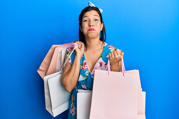 Young latin woman holding shopping bags puffing cheeks with funny face. mouth inflated with air, catching air.  - Foto, imagen