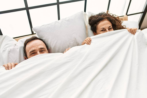 Латиноамериканская пара среднего возраста, покрывающая лицо простыней, лежащих на кровати дома. - Фото, изображение