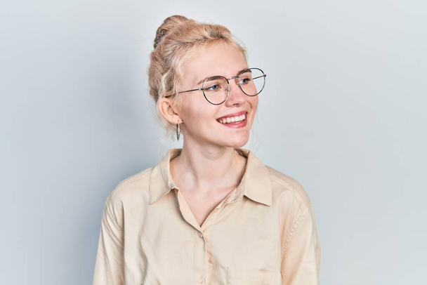 Schöne kaukasische Frau mit blonden Haaren, lässigem Look und Brille, die mit einem Lächeln im Gesicht und natürlichem Ausdruck zur Seite schaut. Lachen selbstbewusst.  - Foto, Bild