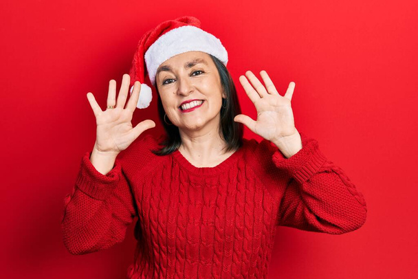 Middelbare leeftijd Spaanse vrouw met kerst hoed tonen en wijzen met vingers nummer tien, terwijl ze vol vertrouwen en gelukkig glimlacht.  - Foto, afbeelding
