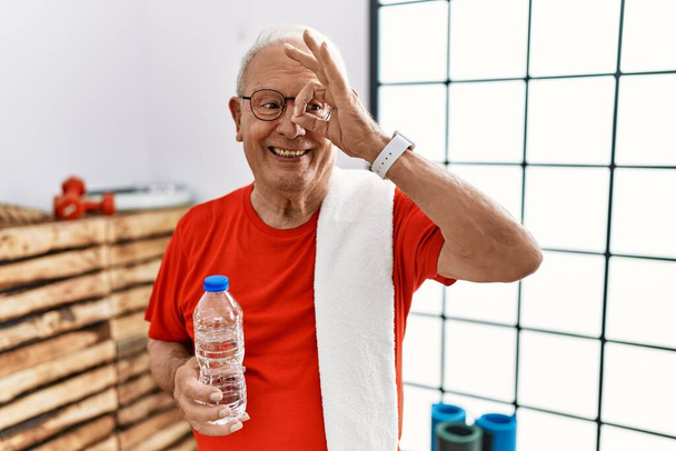 Senior άνθρωπος φορώντας αθλητικά ρούχα και πετσέτα στο γυμναστήριο κάνει εντάξει χειρονομία με το χέρι χαμογελώντας, τα μάτια κοιτάζοντας μέσα από τα δάχτυλα με χαρούμενο πρόσωπο.  - Φωτογραφία, εικόνα