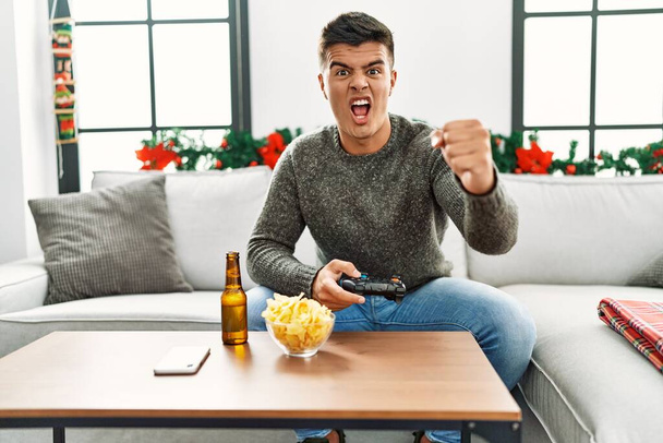 Young hispanic man playing video game on christmas annoyed and frustrated shouting with anger, yelling crazy with anger and hand raised  - Foto, Imagen