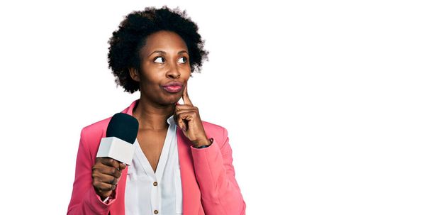 African american woman with afro hair holding reporter microphone serious face thinking about question with hand on chin, thoughtful about confusing idea  - Foto, Imagem