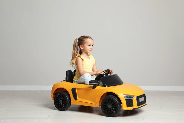 Cute little girl driving children's electric toy car near grey wall indoors - Foto, afbeelding