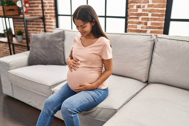 Jovem mulher latina grávida sorrindo confiante sentado no sofá em casa - Foto, Imagem