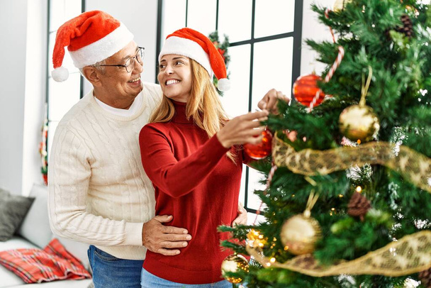 Spanisches Paar mittleren Alters lächelt glücklich beim Schmücken des Weihnachtsbaums zu Hause. - Foto, Bild