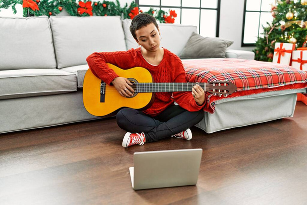 Femme hispanique aux cheveux courts apprenant la guitare classique à partir de tutoriel en ligne attitude de pensée et expression sobre regardant avec confiance en soi  - Photo, image