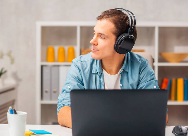 Young man in headphones works on a laptop computer at home. Remote work and education concept. - Foto, imagen