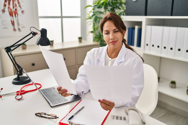 Young latin woman wearing doctor uniform reading paperwork at clinic - Foto, Imagen