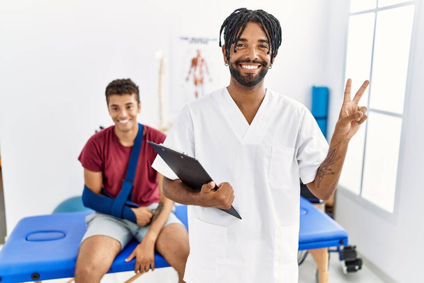 Een jonge Spaanse man die werkt in een pijnkliniek met een man met een gebroken arm die glimlacht met een vrolijk gezicht knipoogt naar de camera en met zijn vingers een overwinningsbord doet. nummer twee.  - Foto, afbeelding