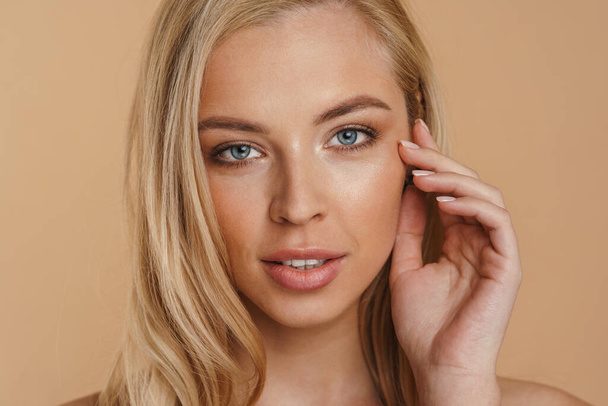 Beauty image of a smiling young blonde woman with long hair naked shoulders standing over beige background touching face - Φωτογραφία, εικόνα