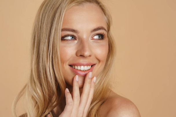 Beauty image of a smiling happy young blonde woman with long hair naked shoulders standing over beige background touching face - Φωτογραφία, εικόνα