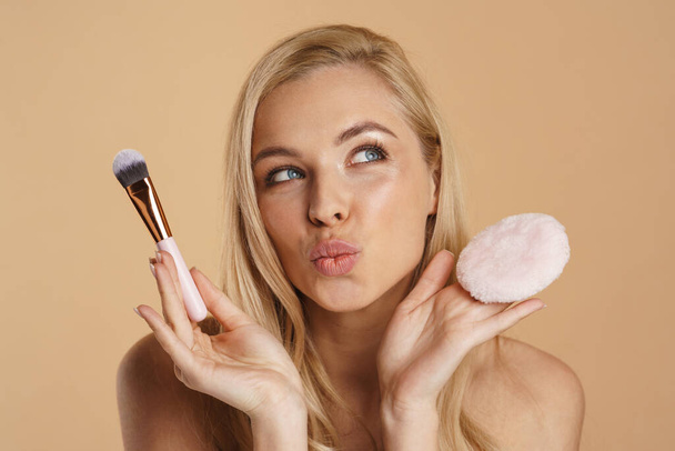 Smiling young blonde woman with long hair with bare shoulders standing over beige background, holding powder puff and makeup brush - Photo, image
