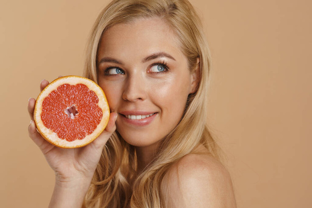 Smiling young blonde topless woman with long hair standing over beige background, holding cut grapefruit - Valokuva, kuva