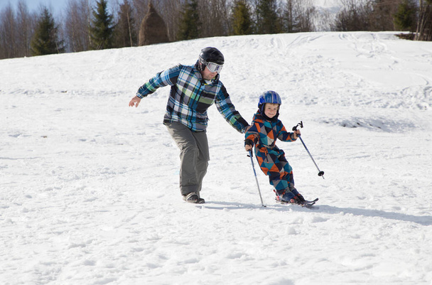 寒い冬の日に雪の山で父とスキーをすることを学ぶ子供の男の子。季節の喜び、幸せな子供時代、健康的なライフスタイル。スポーツ教育、ケア、サポート - 写真・画像