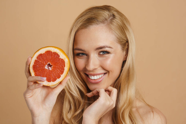 Smiling young blonde topless woman with long hair standing over beige background, holding cut grapefruit - Фото, зображення