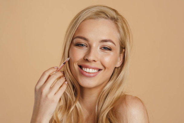 Young white blonde woman with healthy looking skin holding syringe with standing over beige background - Foto, afbeelding